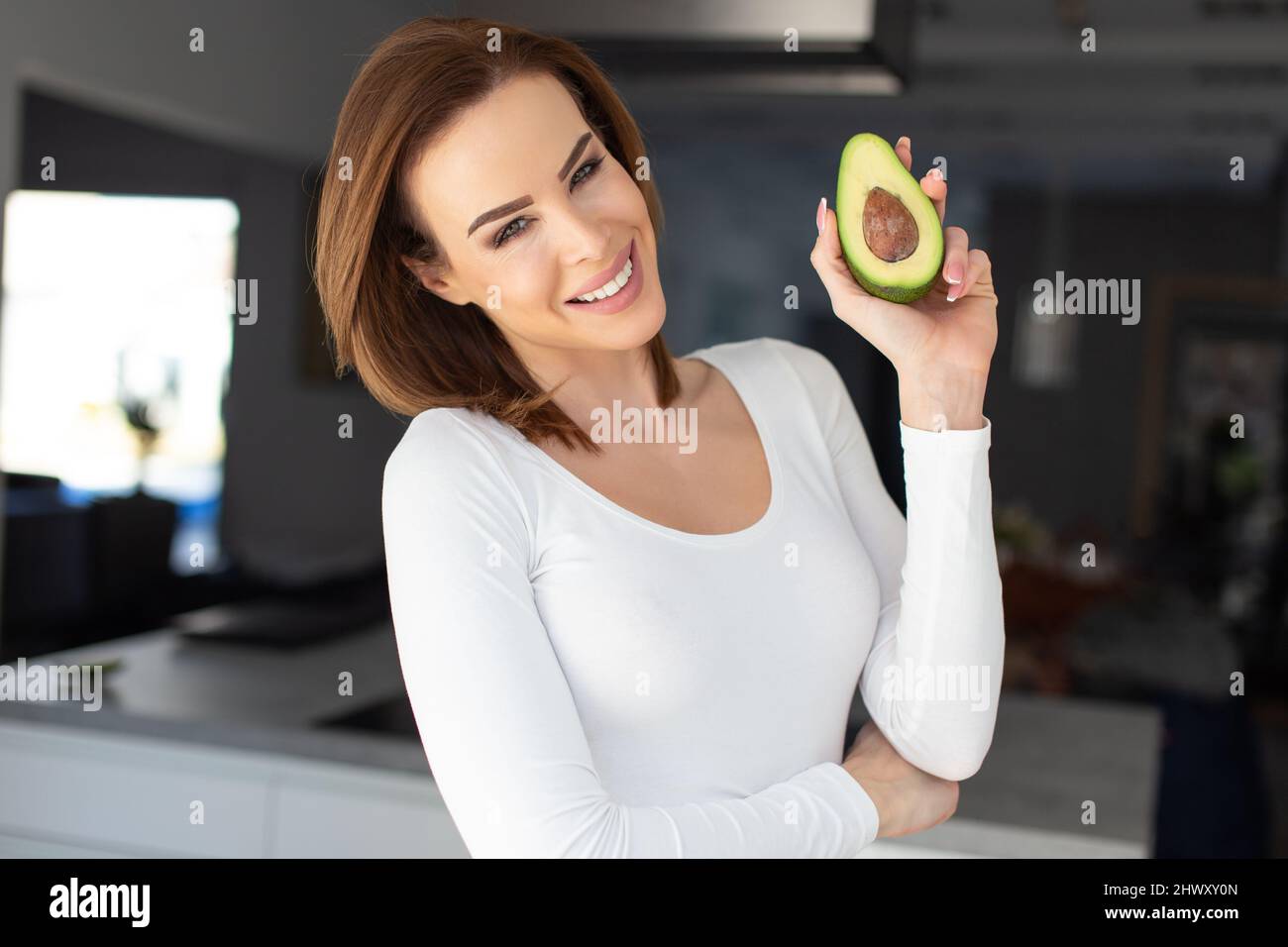 Glückliche junge 30s kaukasische Frau zeigt halbierte Avocado in der Küche, toothy Lächeln Stockfoto