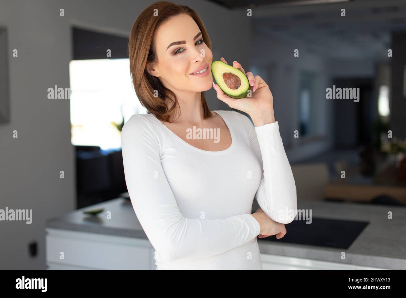 Junge fröhliche 30s kaukasische Frau, die halbierte Avocado im Gesicht in der Küche hält Stockfoto