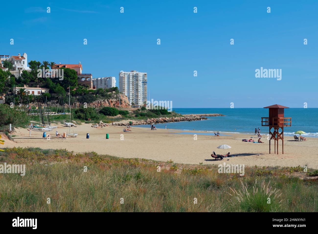 Menschen, die sich mit ihren Sonnenschirmen auf dem Sand sonnen und am Strand von Cap Blanc in der Küstenstadt Cullera in der Provinz Valencia, Spanien, baden, Stockfoto