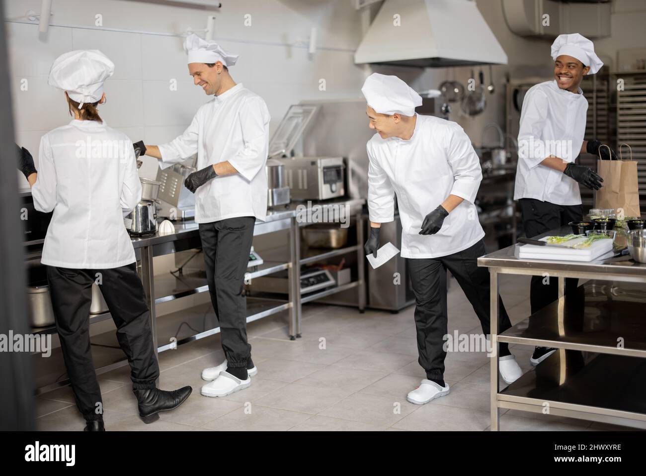 Fröhliches multirassisches Team von Köchen arbeitet aktiv in der Küche. Der asiatische Koch kündigt die Bestellliste vom gedruckten Scheck an, der lateinische Kerl verpackt Lebensmittel für die Lieferung, die europäischen Köche kochen hinterher. Konzept der Teamarbeit im Restaurant Stockfoto