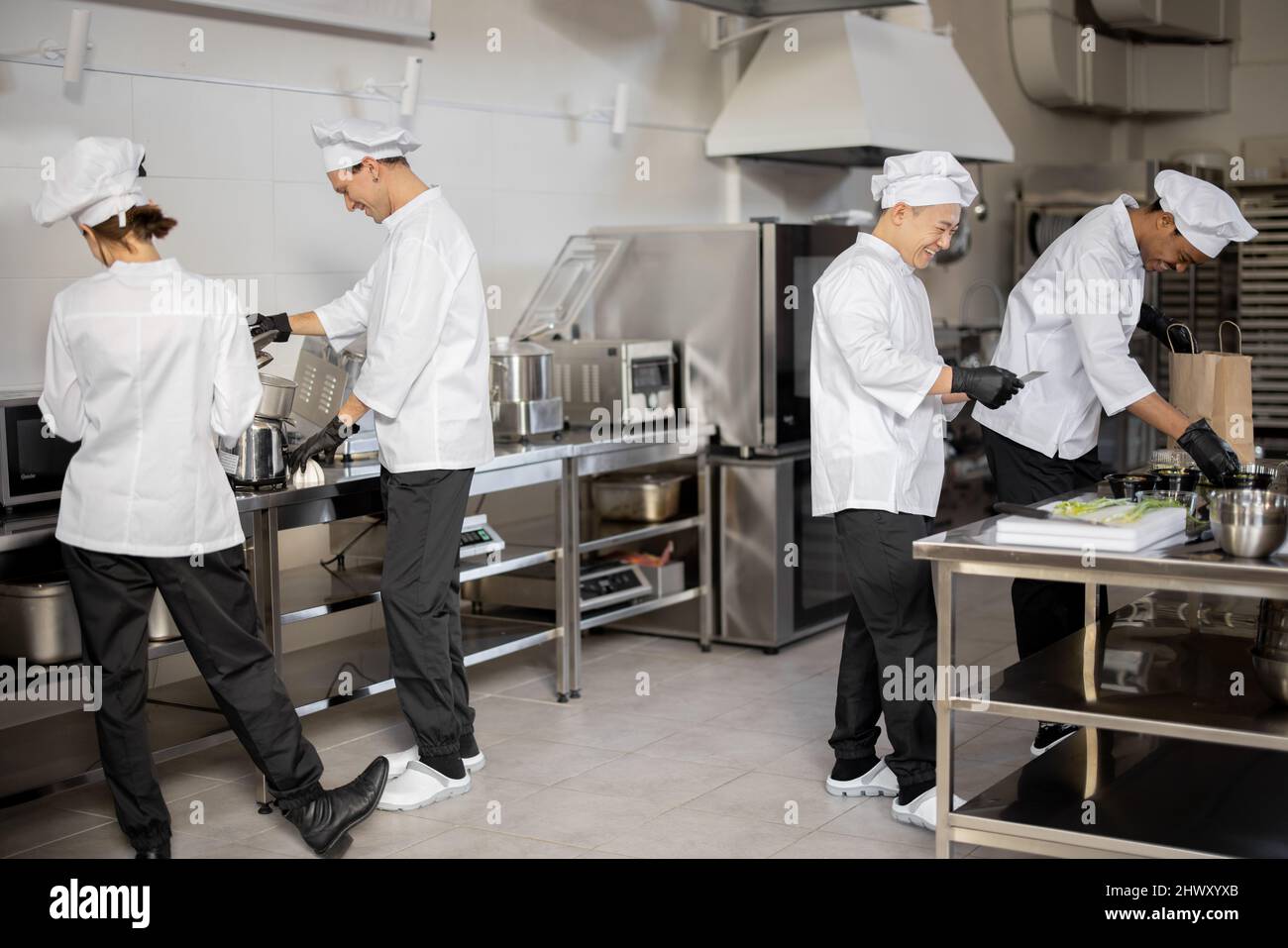 Fröhliches multirassisches Team von Köchen arbeitet aktiv in der Küche. Der asiatische Koch kündigt die Bestellliste vom gedruckten Scheck an, der lateinische Kerl verpackt Lebensmittel für die Lieferung, die europäischen Köche kochen hinterher. Konzept der Teamarbeit im Restaurant Stockfoto