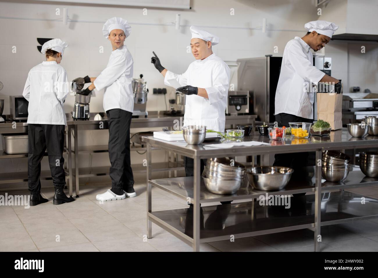 Multirassisches Team von Köchen arbeitet aktiv in der Küche. Der asiatische Koch kündigt die Bestellliste vom gedruckten Scheck an, der lateinische Kerl verpackt Lebensmittel für die Lieferung, die europäischen Köche kochen hinterher. Konzept der Teamarbeit im Restaurant Stockfoto
