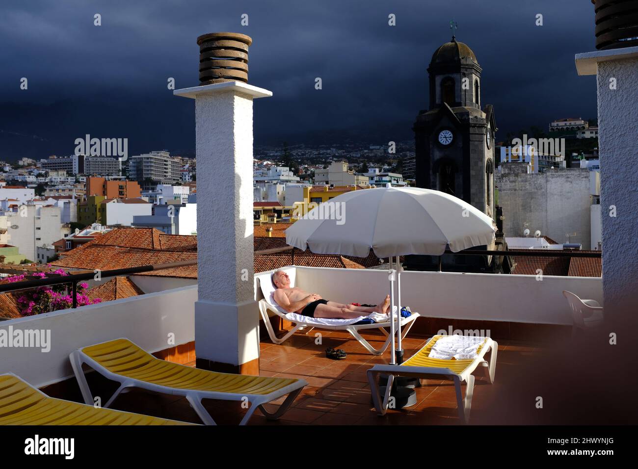 Ein älteres Männchen, das auf einem Sonnenbett mit der Stadt Puerto de la Cruz, Teneriffa, im Hintergrund, Stockfoto