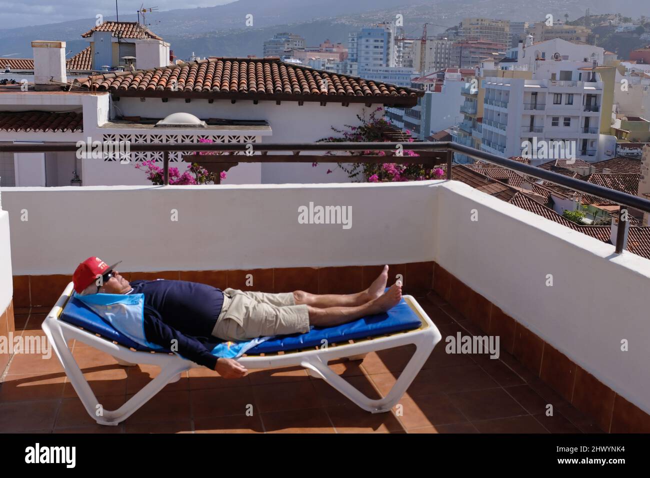 Ein älteres Männchen, das auf einem Sonnenbett mit der Stadt Puerto de la Cruz, Teneriffa, im Hintergrund, Stockfoto
