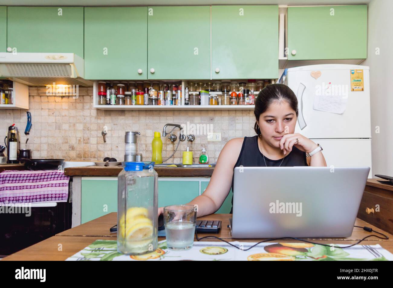 Vorderansicht einer jungen lateinischen kaukasischen Frau, die mit ihrem Computer von zu Hause aus studiert, Online-Kurse besucht und mit einer Hand auf den Computer starrt Stockfoto