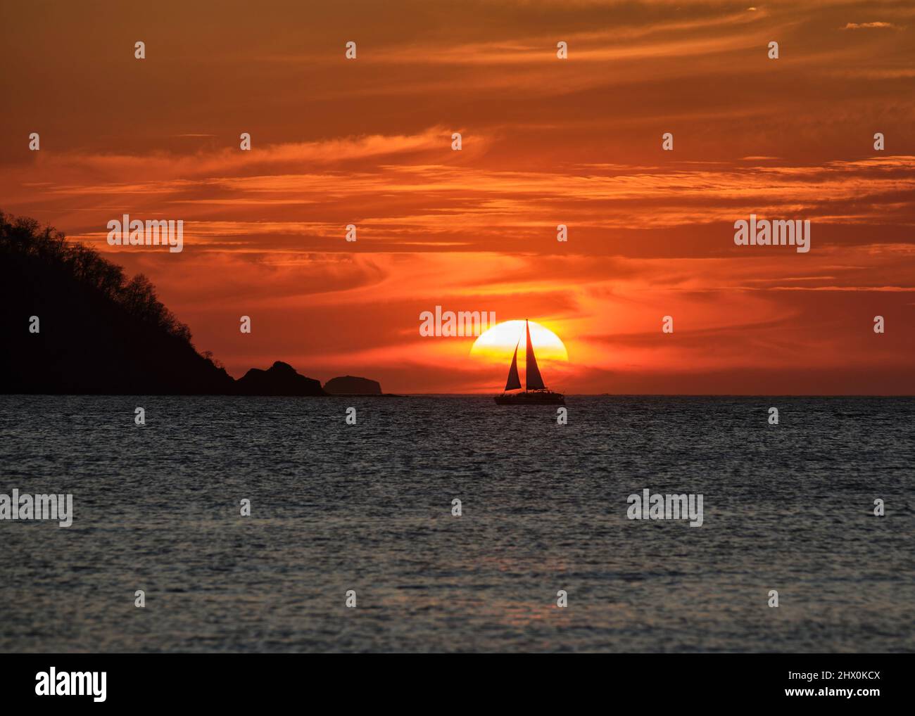 Ein spektakulärer orangefarbener Sonnenuntergang mit wirbelnden Wolken und der Silhouette eines Segelbootes vor der Sonne in Potrero Costa Rica. Stockfoto