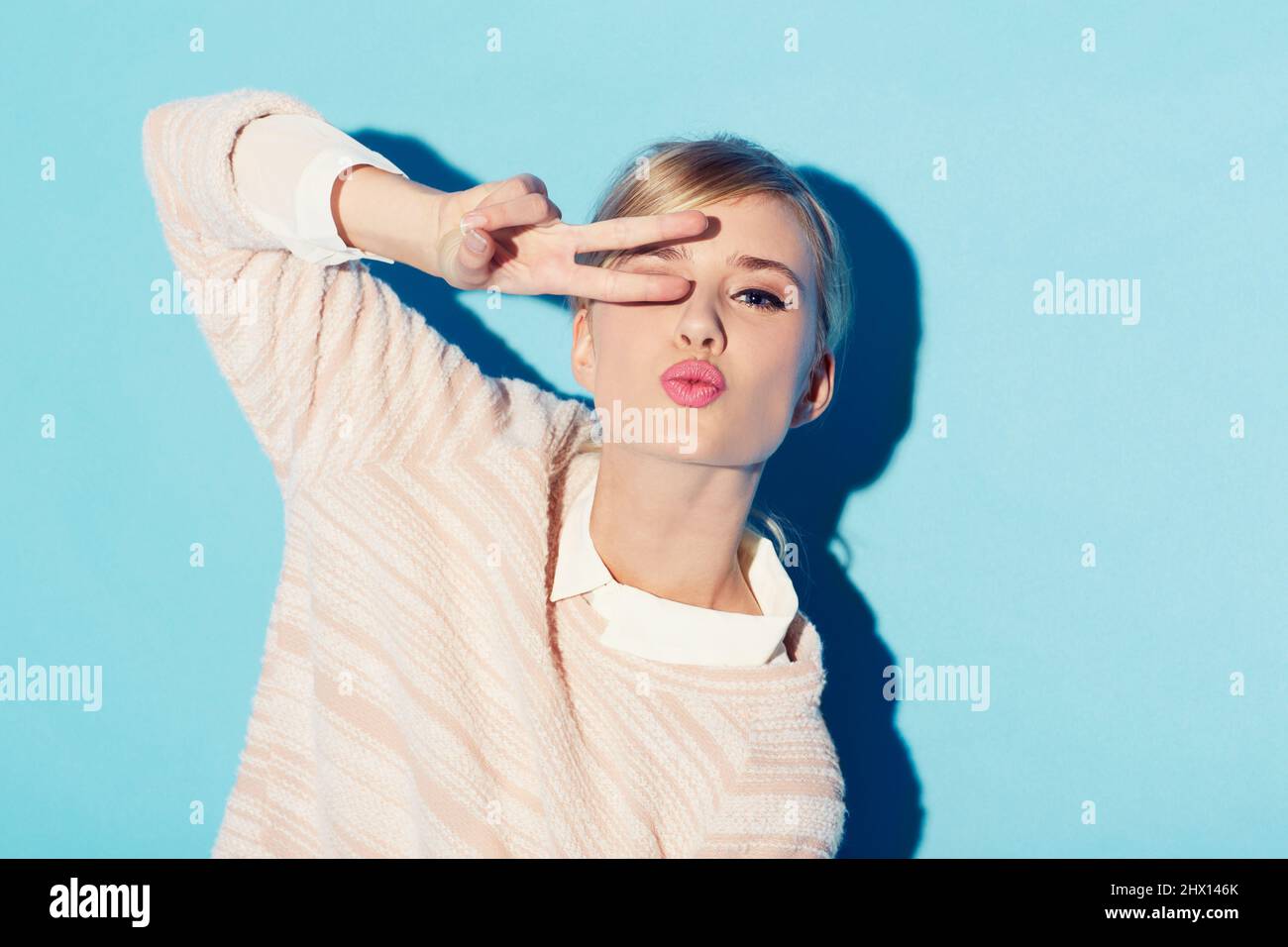 Frieden, Liebe und Schönheit. Eine wunderschöne junge Frau bedeckt ihr ein Auge mit zwei Fingern, während sie vor einem blauen Hintergrund steht. Stockfoto