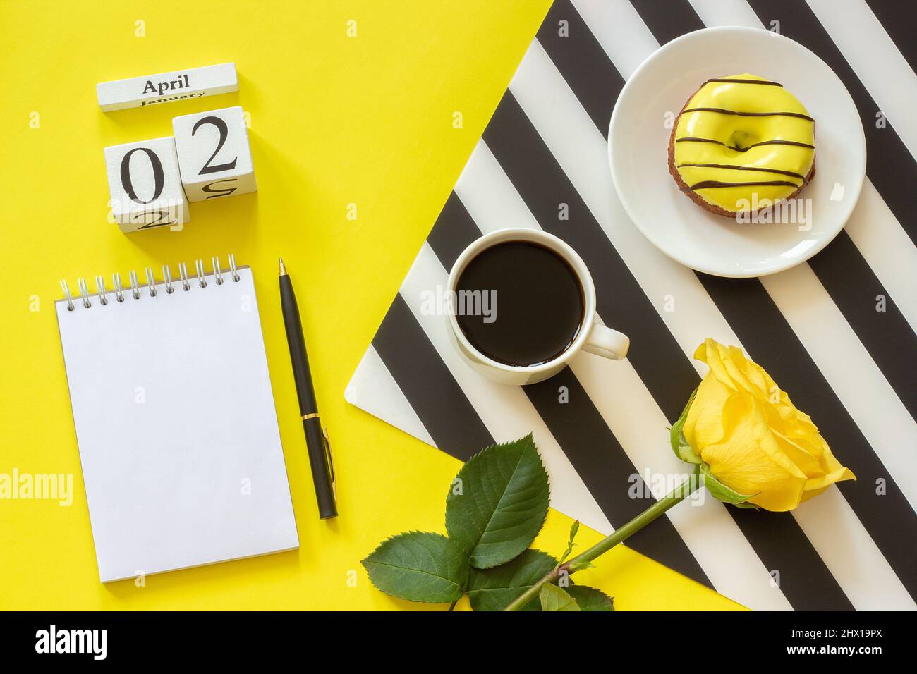 Holzwürfel Kalender April 2.. Tasse Kaffee, gelber Donut und Rose auf schwarz-weißer Serviette, leerer offener Notizblock für Text auf gelbem Hintergrund. Kontra Stockfoto