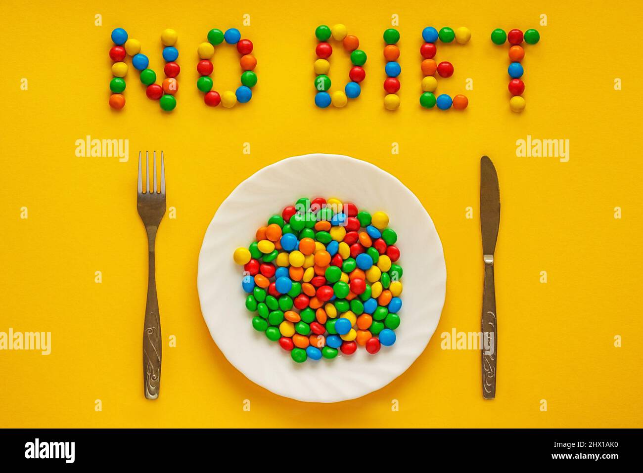 Bunte Bonbons auf Teller und Besteck (Messer und Gabel) auf gelbem Hintergrund. Inschrift „Keine Diät“. Concept International No Diet Day ist feiern Stockfoto