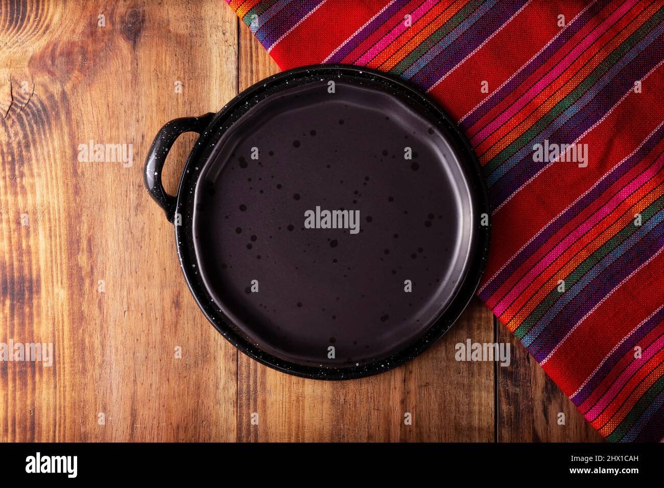 Mexikanische Küchenutensilien, bunte traditionelle Stoffe, leere schwarze Platte auf comal de peltre auf rustikalem Holztisch. Flatlay Stockfoto