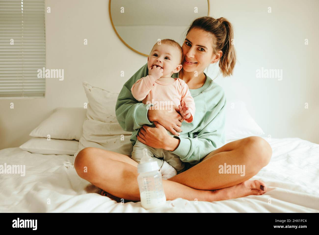 Glückliche Mutter lächelt der Kamera zu, während sie ihr entzückendes Baby hält. Fröhliche junge Mutter, die mit ihrem neugeborenen Baby auf dem Bett sitzt. Fürsorgliche Mutter Kreati Stockfoto