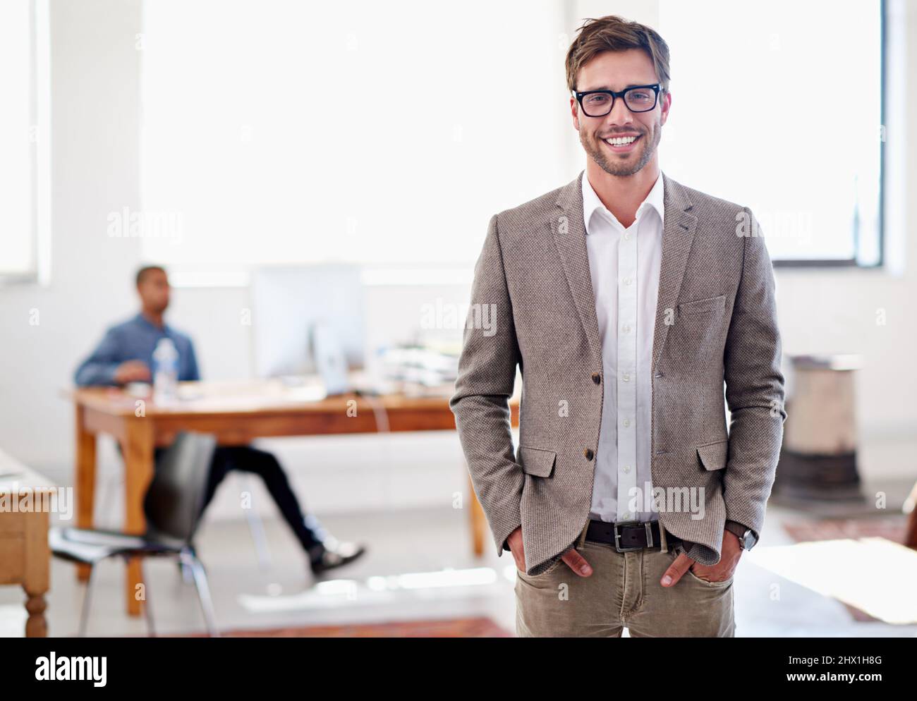 Sie werden dieses Büro lieben. Porträt eines jungen Mannes, der in einem Büro mit einem Mitarbeiter im Hintergrund steht. Stockfoto