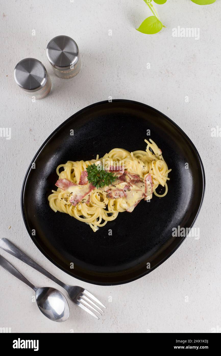 Eine gekochte Pasta-Spaghetti Carbonara mit Sahnesauce, Rindfleisch, Olivenöl und frischer grüner Petersilie in einem schwarzen ovalen Teller mit einem Paar Bestecke Stockfoto