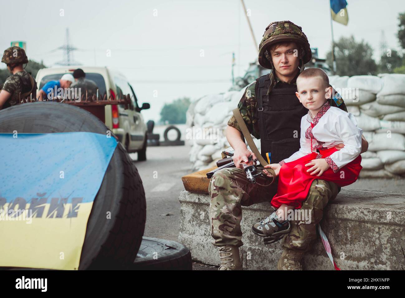Ukrainischer Junge in traditioneller Nationaltracht sitzt in der Nähe eines ukrainischen Soldaten auf einer Straßensperre vor dem Hintergrund von Sandsäcken. Konzept des russischen Militärs Stockfoto