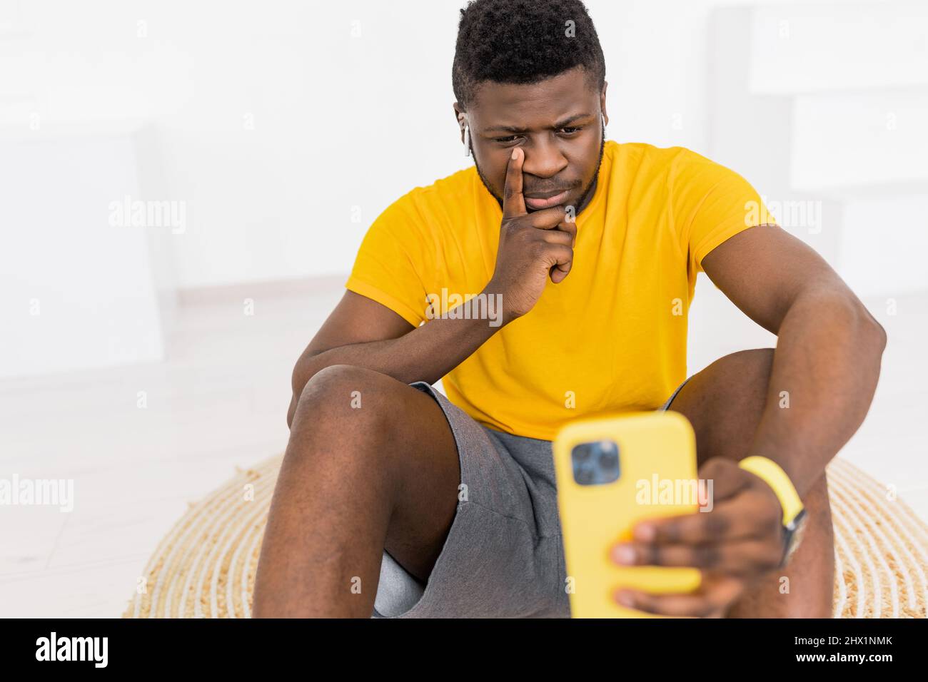 Schöner afroamerikanischer schwarzer Mann ernst zu gelb Smartphone mit kabellosen Kopfhörern. Selbstporträt. Aufgeregter junger afrikanischer Mann in Gelb Stockfoto