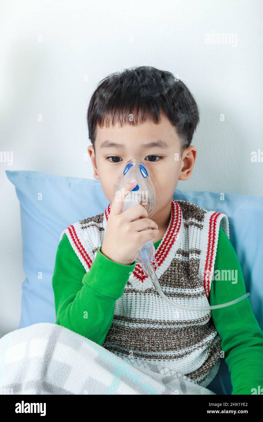 Trauriges asiatisches Kind hält einen Maskendampf-Inhalator zur Behandlung von Asthma auf Krankenbett im Krankenhaus. Atmen durch einen Dampfvernebler. Konzept der Inhalation Stockfoto