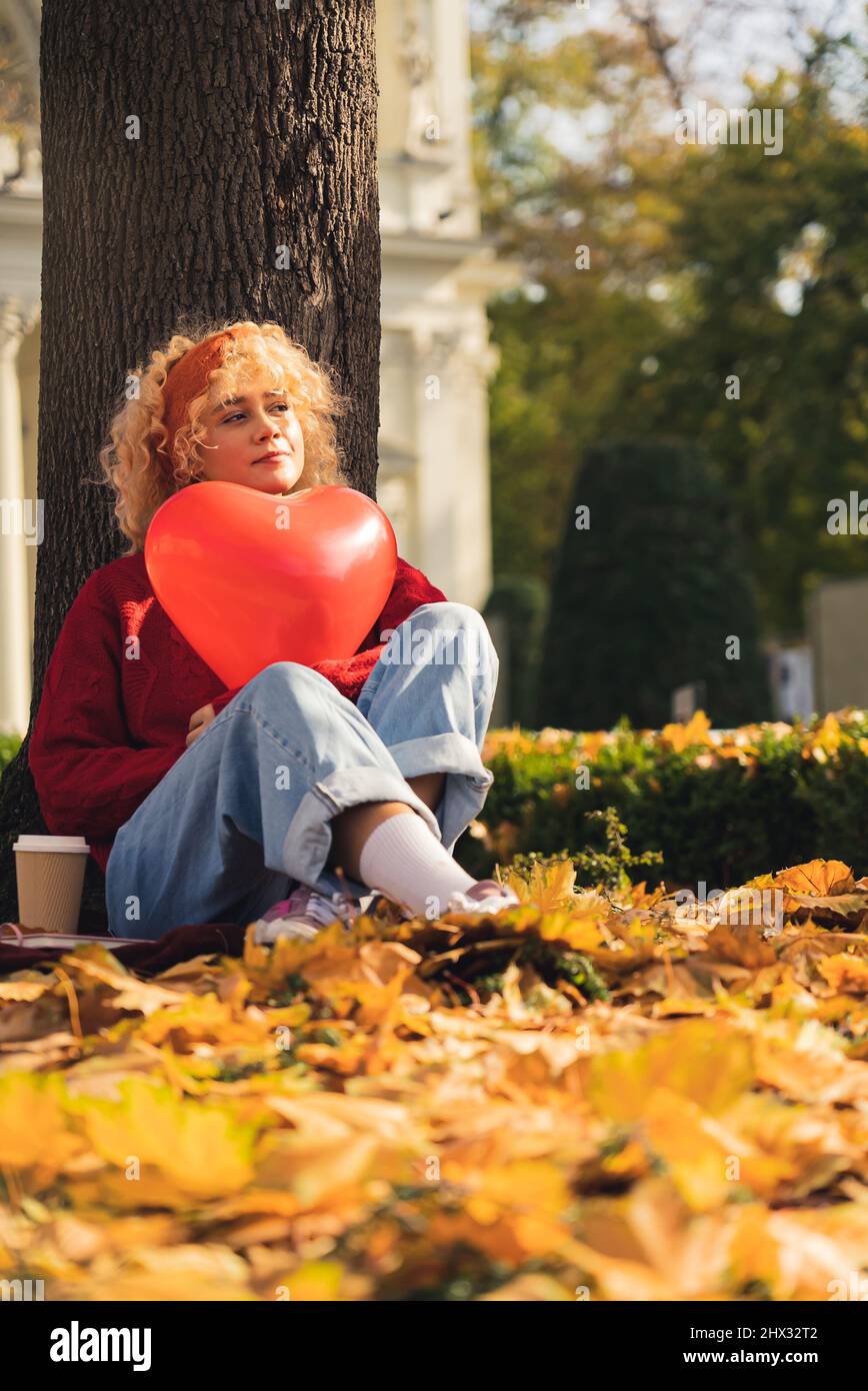 Nachdenkliche junge Frau, die unter den Herbstblättern des Baumes sitzt und einen roten Herzballon hält - Gesamtaufnahme. Hochwertige Fotos Stockfoto