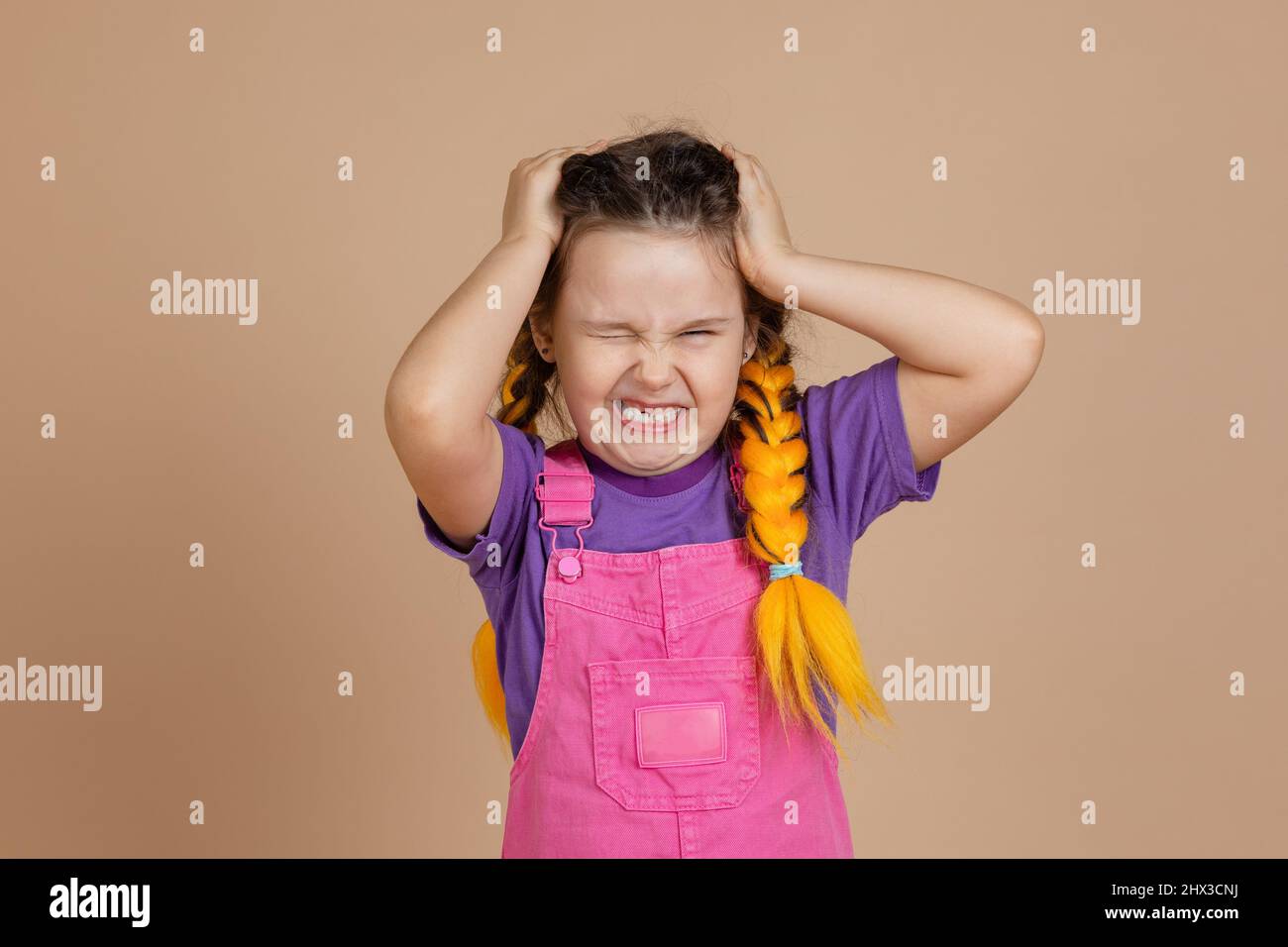 Irritiertes kleines Mädchen, das Kopfschmerzen hat, den Kopf berührt, wobei die Hände gelbe Kanekalon-Zöpfe haben, die in rosa Jumpsuit und violettem T-Shirt grinsend sind Stockfoto