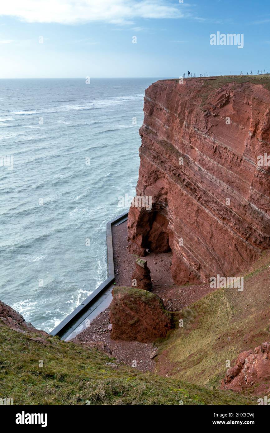 Menschen stehen am Rande der hohen roten dramatischen Klippen der Insel Helgoland mit rauem Meer. Sonniger windiger Wintertag in Helgoland an der Nordsee Stockfoto