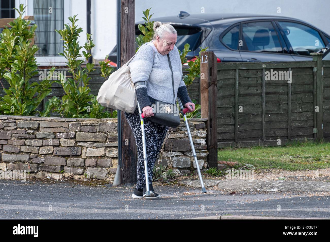 Ältere Frau, die mit Krücken die Straße entlang läuft und eine Einkaufstasche trägt, Großbritannien. Thema - Behinderung, Mobilitätsprobleme Stockfoto