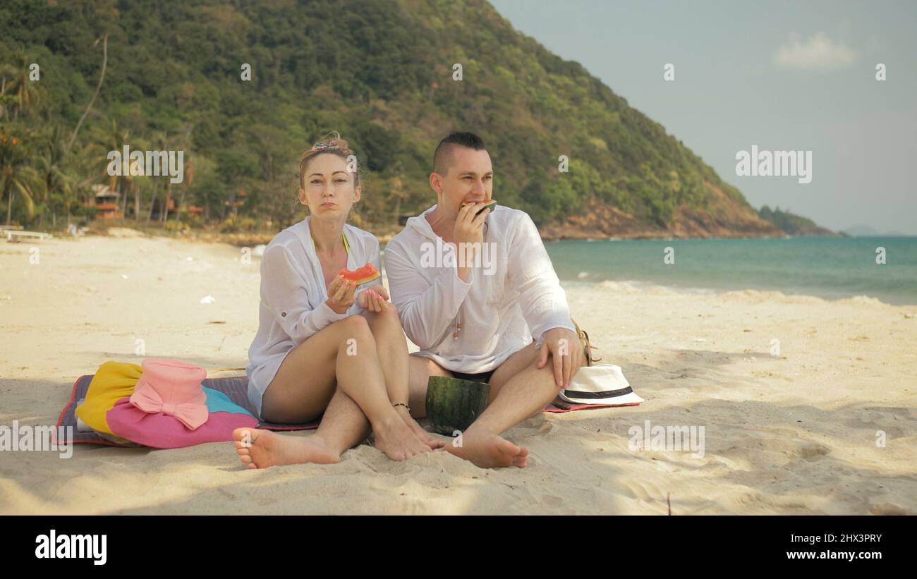 Das fröhliche Liebespaar hält und isst Wassermelonenscheiben am tropischen Sandstrand. Romantische Liebhaber zwei kaukasische Menschen verbringen das Sommerwochenende im Freien. Mütze, Rucksack weißes Hemd Strandmode. Stockfoto
