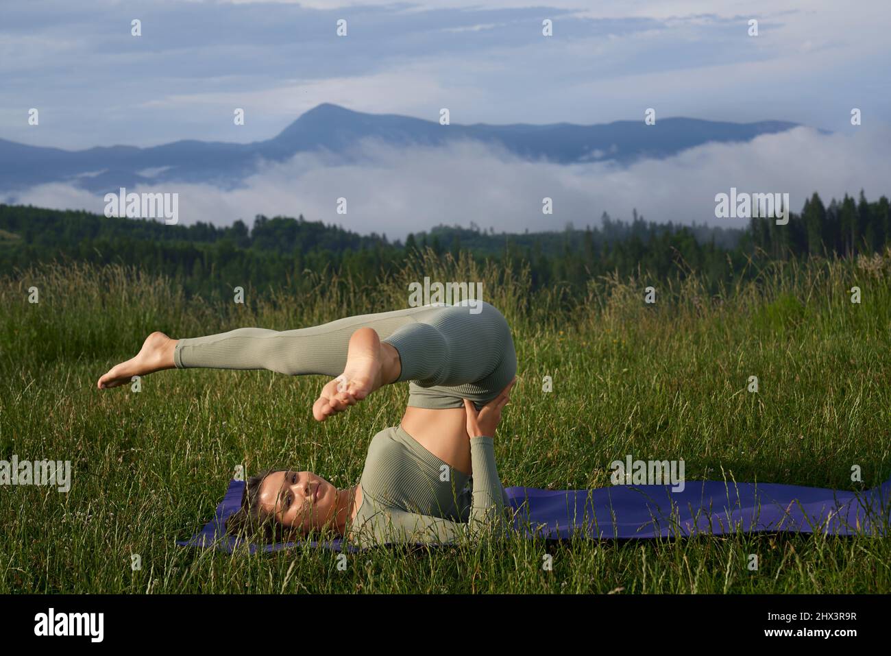 Sportliche junge Frau in grüner aktiver Kleidung, die auf Matte unter grünen Bergen Akro-Yoga macht. Hübsche Dame, die sich durch regelmäßiges Training an der frischen Luft um den Körper kümmert. Stockfoto