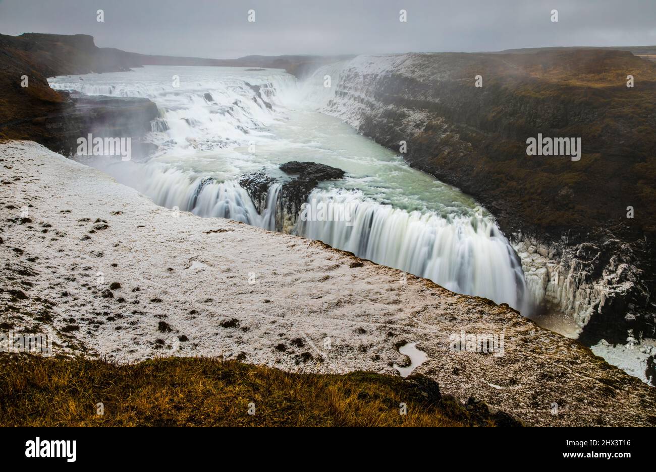 Landschaft eines Hügels mit einem Fluss und dampfenden Wasserfällen in Island unter bewölktem Himmel. Stockfoto