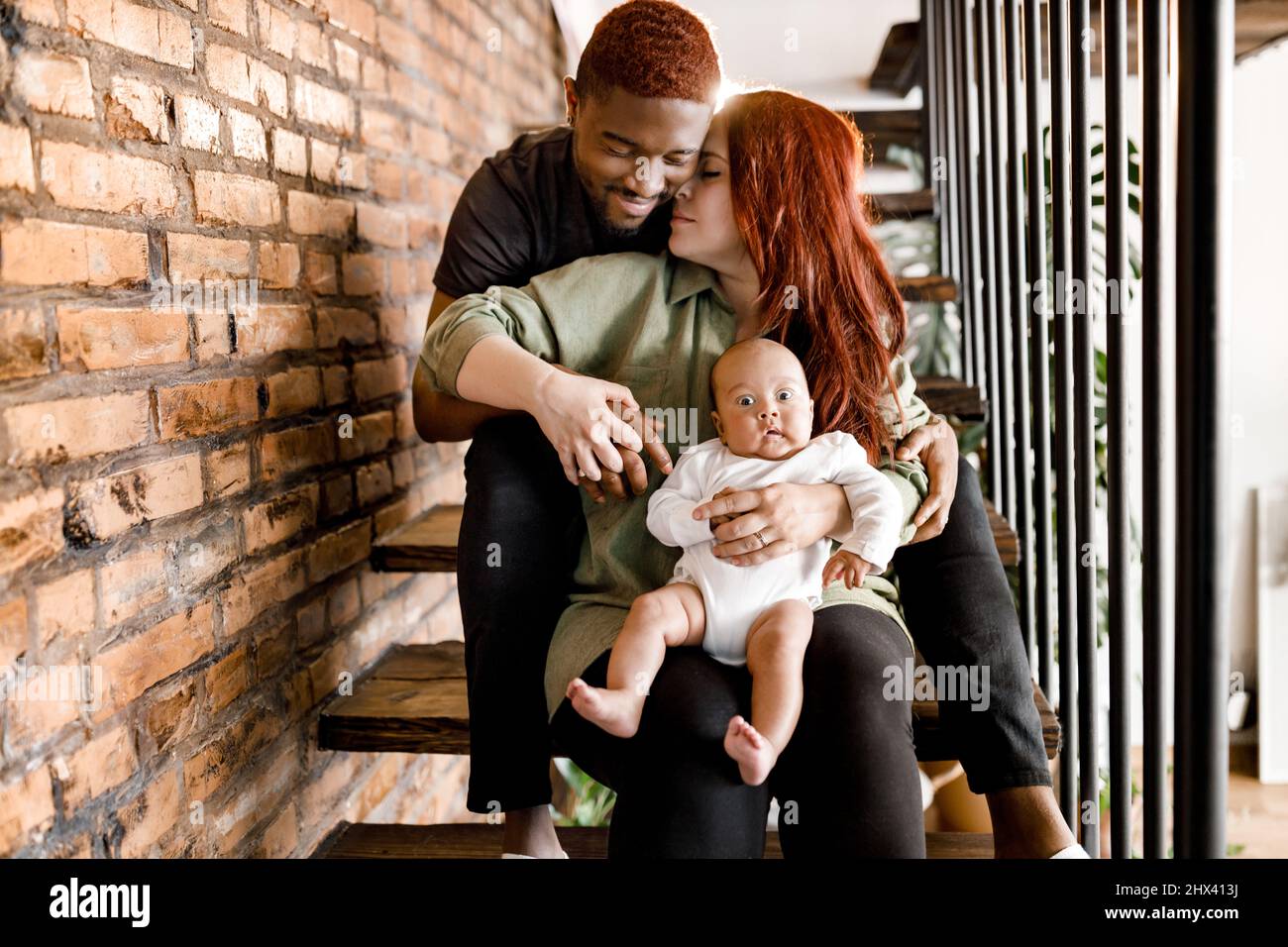 Porträt einer gemischten Rasse lächelnde Familie sitzt auf der Treppe. Schöne Mutter und lächelnder Vater hält Baby Junge. Fröhliche Eltern schauen sanft auf ihre Stockfoto