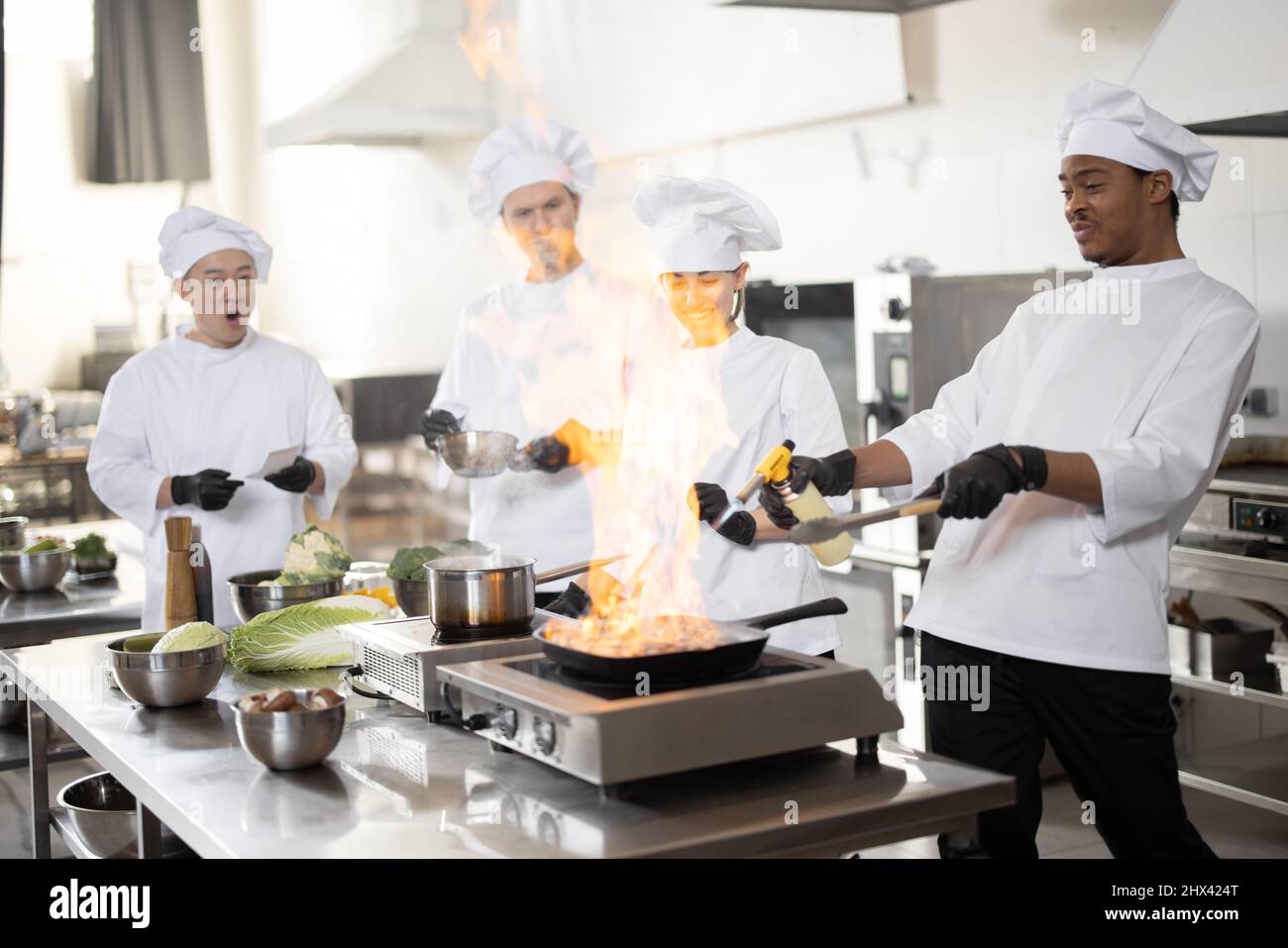 Multirassisches Team professioneller Köche in Uniform, die Mahlzeiten für ein Restaurant in der Küche zubereiten. Latein Burning Pan, europäische Köche machen Sauce und asiatischen Koch verwalten den Prozess. Teamarbeit und harter Job im Restaurant Stockfoto