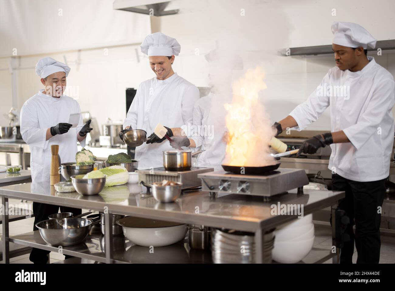 Multirassisches Team professioneller Köche in Uniform, die Mahlzeiten für ein Restaurant in der Küche zubereiten. Latein Burning Pan, europäische Köche machen Sauce und asiatischen Koch verwalten den Prozess. Teamarbeit und harter Job im Restaurant Stockfoto