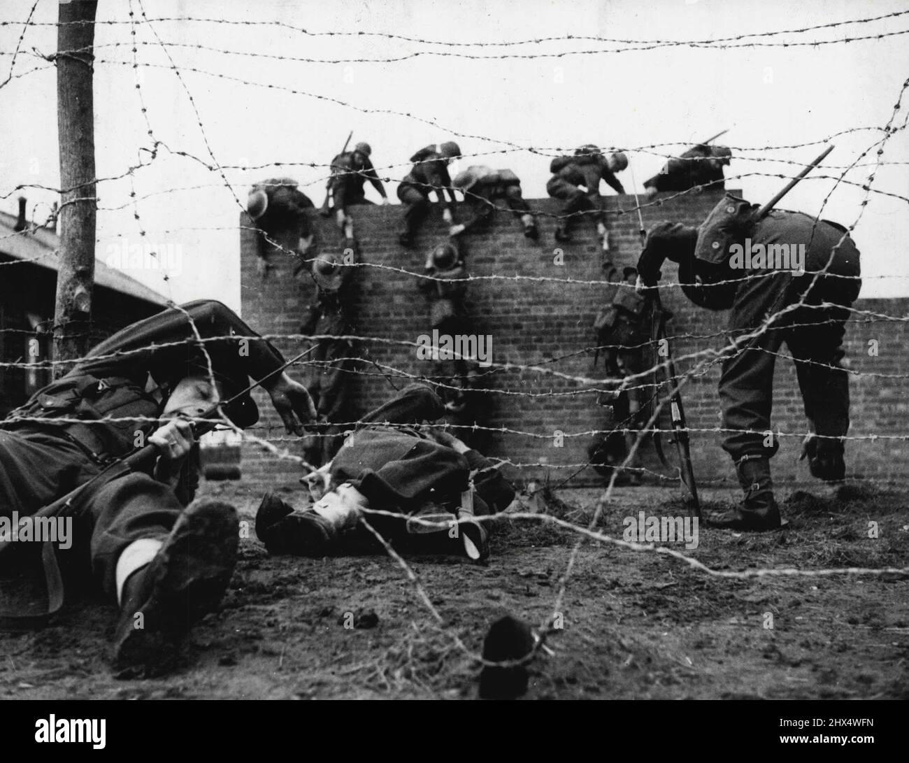 Der „Härtung“-Angriffskurs der Armee und Härtungs-Übungen – das Bild zeigt, wie man durch ein Stacheldraht-Hindernis kommt, während andere Männer über eine zehn Fuß lange Wand hinausragen. Bilder, die die Schwere der Verhärtung der Ausbildung von Armeepersonal auf einem modernen Angriffskurs zeigen, wo neue Methoden eingeführt wurden. Alle Klassen von Armeepersonal - Infanterie, Artillerie, medizinische Einheiten, Panzerbesatzungen, Signale, Nehmen Sie den Kurs. 24. März 1942. (Foto von einem offiziellen britischen Foto). Stockfoto