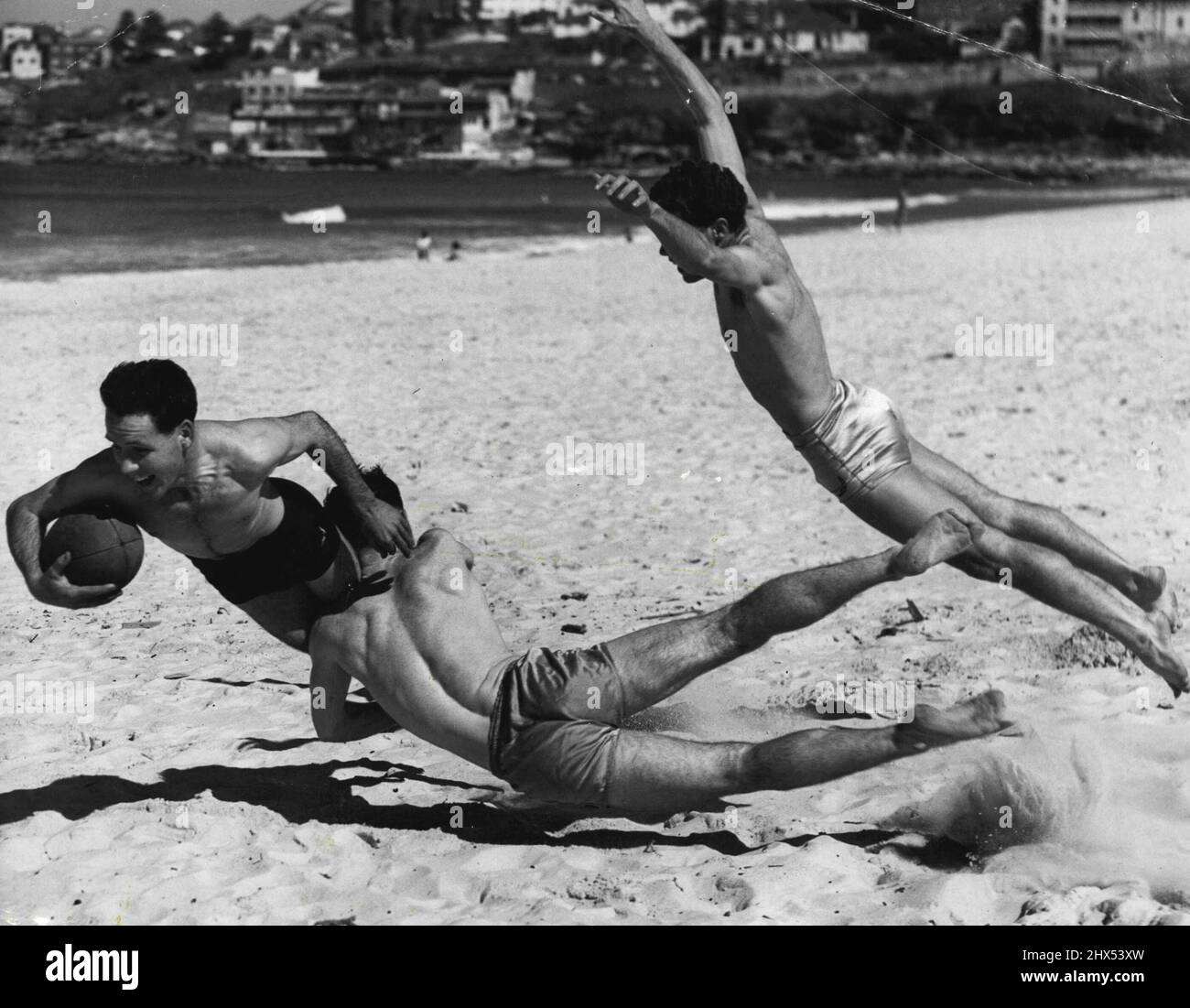 Um sich warm zu halten, haben diese drei Mitglieder des Drummoyne Rugby Union Club heute am Bondi Beach ein Fußballspiel gespielt. Johnny Denton von Ryde wird von John Stephenson von Ryde und Ian Findlay von Filvedock angegangen. 28. August 1955. (Foto von Ronald Leslie Stewart/Fairfax Media). Stockfoto