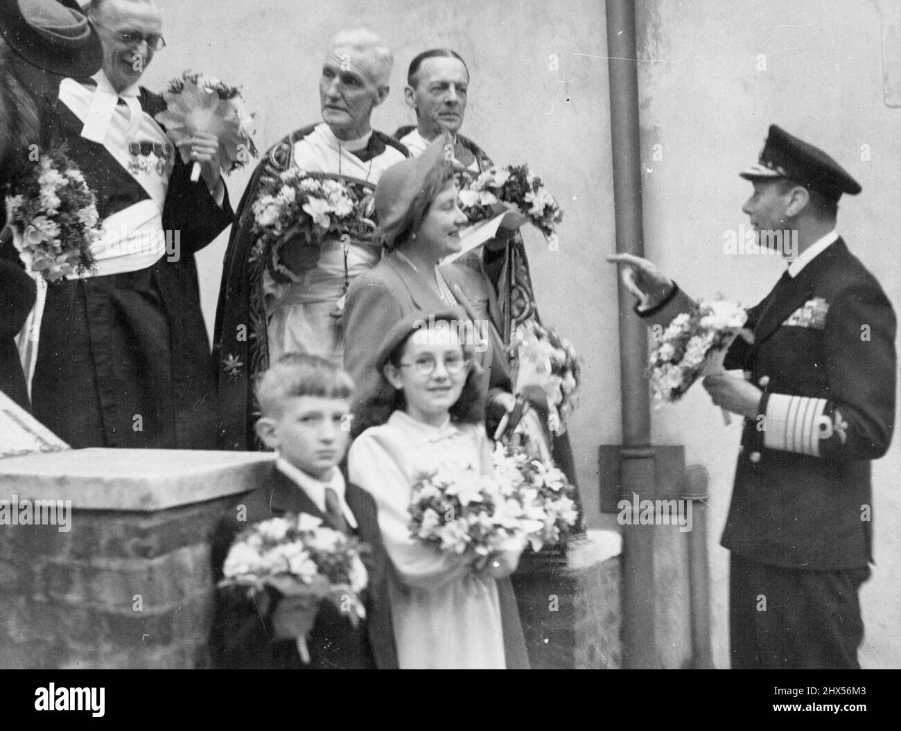 Der König mit Posies, arrangiert ein Gründungstag-Bild -- ein informelles Foto des Königs, trägt einen Blumenpfosten, arrangiert eine Gruppe für ein Foto nach der Gründungstag-Zeremonie bei We ***** Abbey heute Apr 18. Hinter der Königin steht der Bischof von Lichfield, der den Gründunggottesdienst leitete. Der König verteilte heute, am 18. April, das Royal Maundy Geld in der Westminster Abbey. 05.Mai 1946. (Foto von Associated Press Photo) Stockfoto