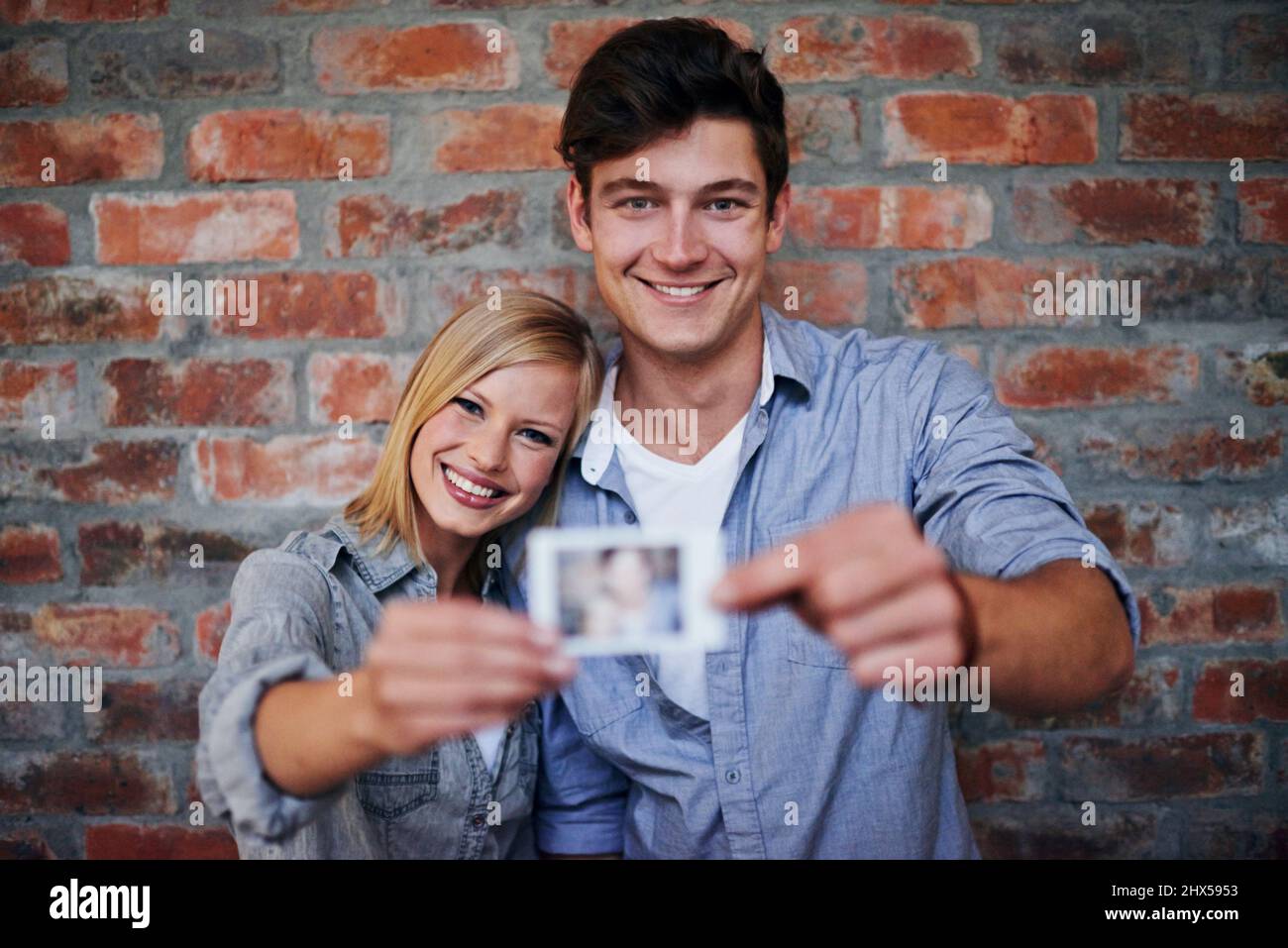 Gemeinsam Erinnerungen schaffen. Ein junges Paar, das vor einer Ziegelwand steht und ein Foto an die Kamera hält. Stockfoto