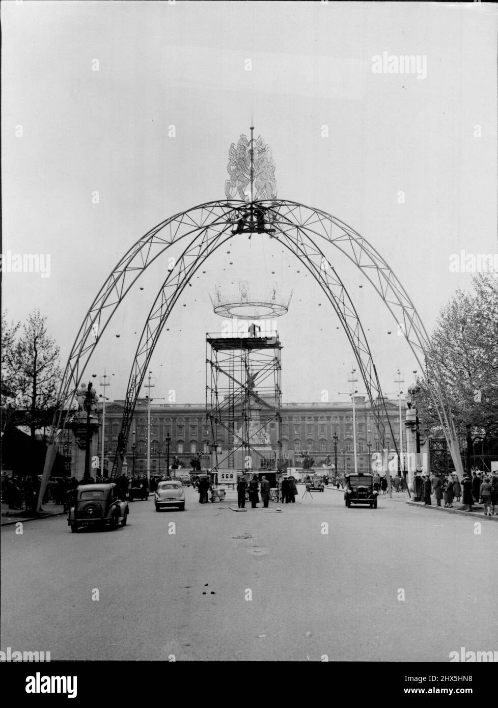 Die Mall Bekommt Diesen Krönungslook -- Auf den Bürgersteigen versammelten Massen und mit dem Buckingham Palace als Rückentuch heben Arbeiter eine Replik einer Königskrone, die an einem 65 Fuß hohen Bogen hängt - einer von vier, die Teil der Krönungsdekorationen in der Mall, London, sein werden. Auf dem Bogen stehen ein Löwe und ein Einhorn, in Stahlrohren. Die Krone wurde heute (Samstag) in Position gebracht. 16.Mai 1953. (Foto von Reuterphoto). Stockfoto