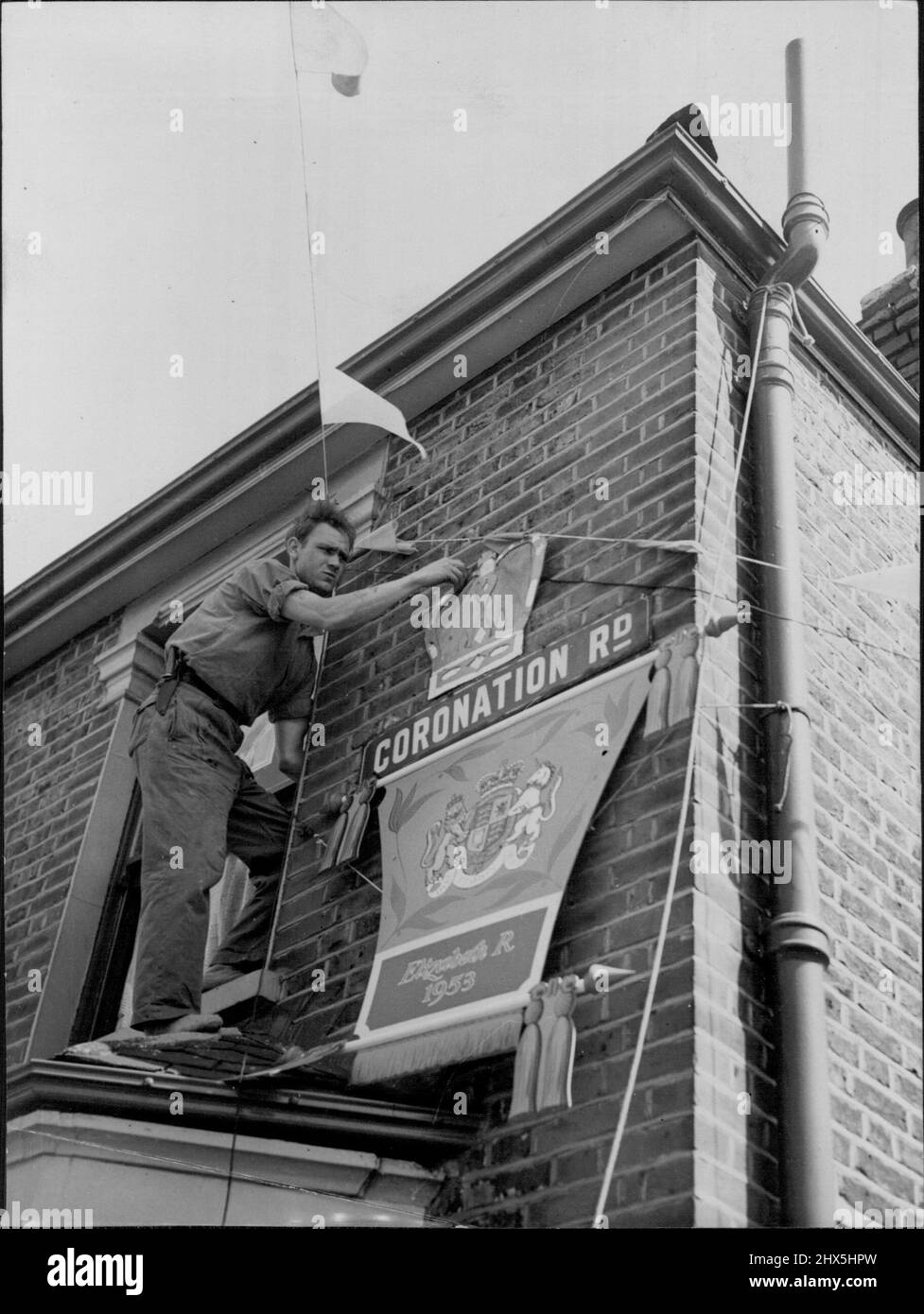 Loyale Grüße - von der Krönungsstraße -- gefährlich auf einem Fenster thront - ein Bewohner der Krönungsstraße (im Stadtteil West Ham, London) fixiert Krönungsdekorationen in Vorbereitung auf den großen Tag. Dies war das Geschenk eines Geschäftsmannes im West End, der nach einer Krönungsstraße suchte - und sie in West Ham fand. Er bot alle notwendigen Verpacke an, um die Straße zu schmücken. 29.Mai 1953. (Foto von Reuterphoto). Stockfoto