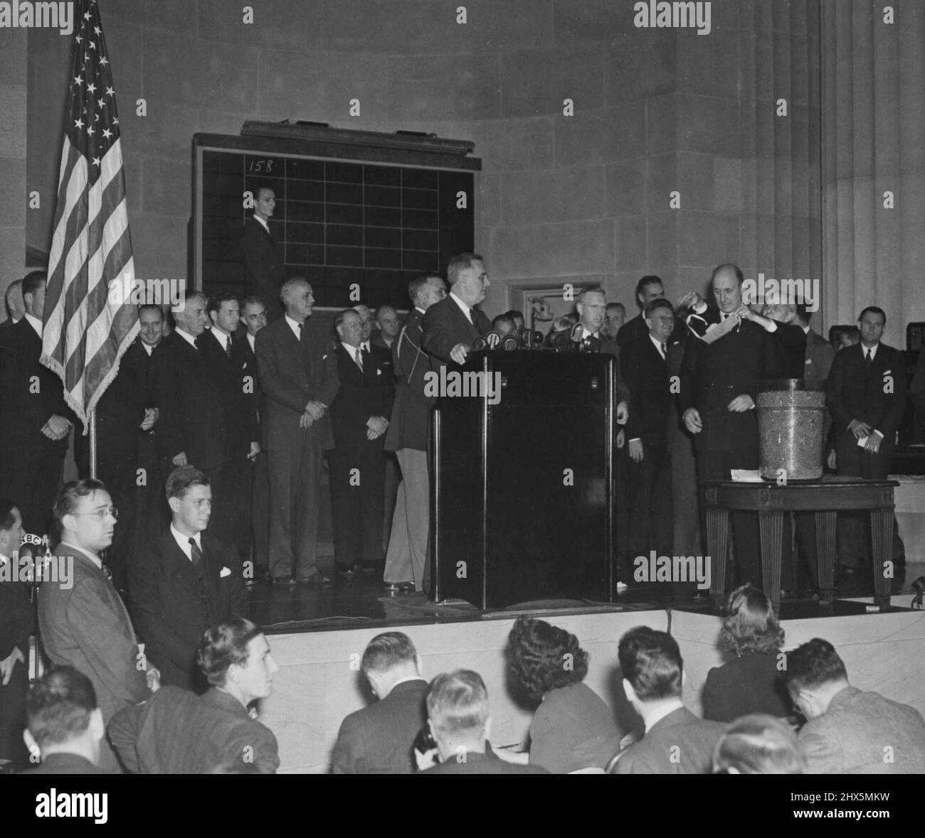 Sekretär Morganthau zieht Nummer -- Finanzminister Henry Morganthau, Jr., der mit verbundenen Augen vor dem Ziehen der zweiten Nummer des Entwurfs der Lotterie in Washington am 29.. Oktober gehalten wird. Colonel Charles R. Morris wendet die Augenbinde an, während Präsident Franklin D. Roosevelt vom Rednerstand aus auf die Seite schaut. Die erste gezeichnete Zahl, 158, ist auf der Tafel im Hintergrund gepostet. 29. Oktober 1940. (Foto von ACME). Stockfoto