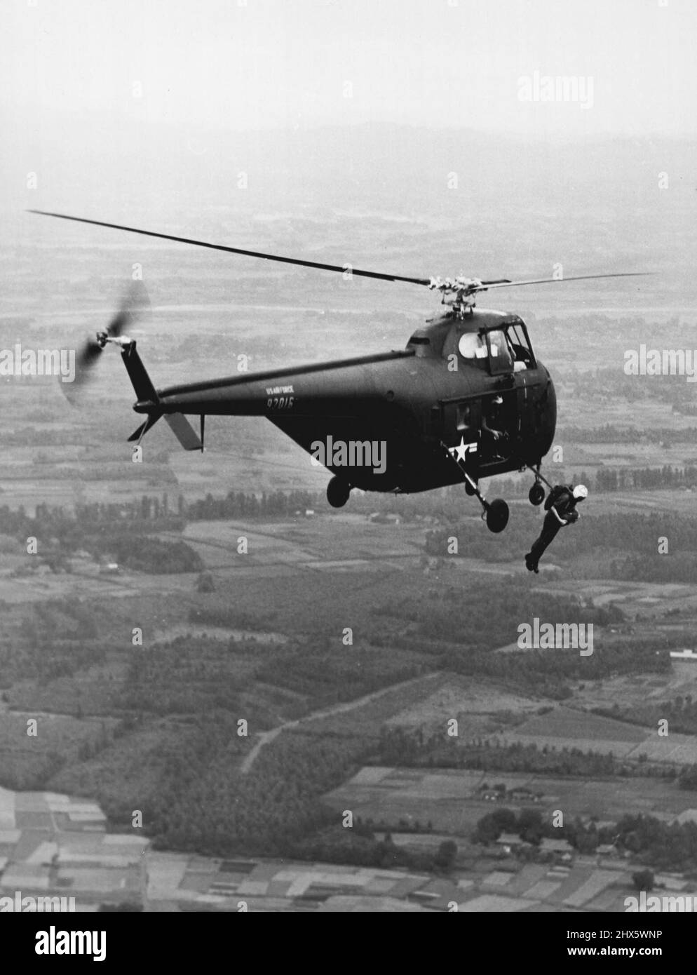 Machen Sie den ersten Sprung von H-19 -- ein Luftwaffenstützpunkt in Japan -- Dieses dramatische Luft-Luft-Foto zeigt einen der vier Männer des 3. Air Res­cue Squadron para-Rescue Teams der US Air Force, der zuerst mit den Füßen eines H-19 Sikorsky Hubschraubers auftaucht. Dies war eine Trainingsmission, die in der Nähe des Heimatstützpunktes der Einheit in Japan stattfand, um die Machbarkeit der Verwendung des größeren „Copters in Fallschirmabschirmmoperationen“ zu testen. Major John C. Shumate Little Rock, Ark, nahm an dem historischen Sprung Teil. Stockfoto