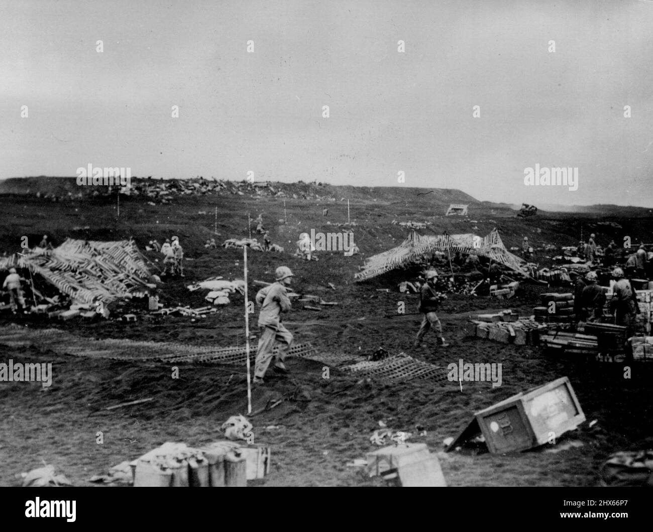 'Rush Job' -- während Foxholes und Gewehrplatzierungen in Eile im 'Hintergrund' getarnt werden, legen Elemente der Fifth Marine Division Stahlmatten über weiche vulkanische Asche des Iwo Jima Beach, um ein Straßenbett zu bilden, um eine Müllkippe auf der rechten Seite zu liefern. Es wurden bereits Vorräte von Marine- und Küstenwache-bemannten Landungsschiffen an Land gebracht, um die Arbeit am Gang zu halten. 7. März 1945. (Foto vom offiziellen Foto der US-Küstenwache). Stockfoto