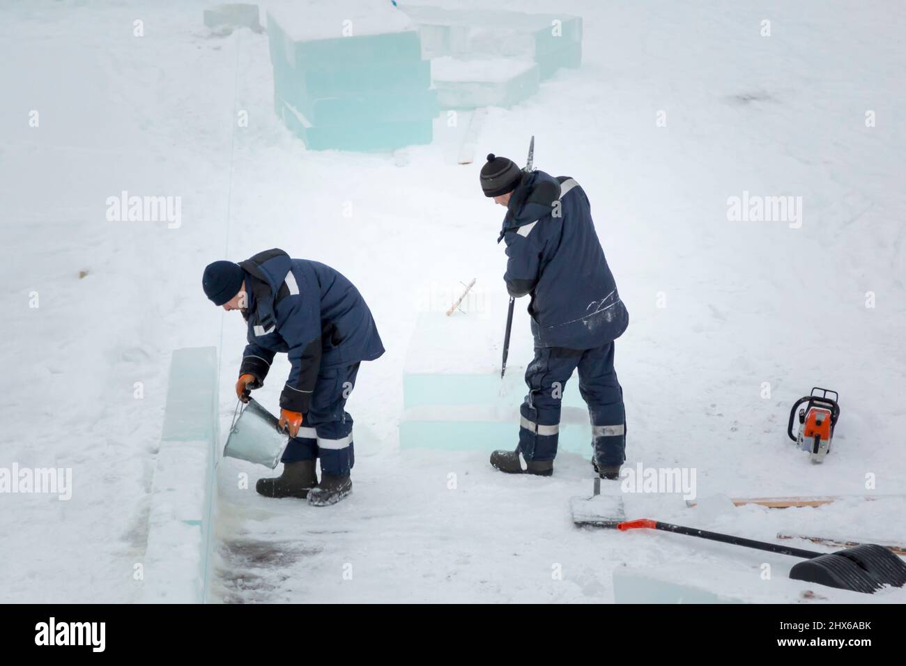 Ein Eisstädter in einer blauen Kapuzenjacke gießt Wasser aus einem Eimer über einen Eisblock Stockfoto