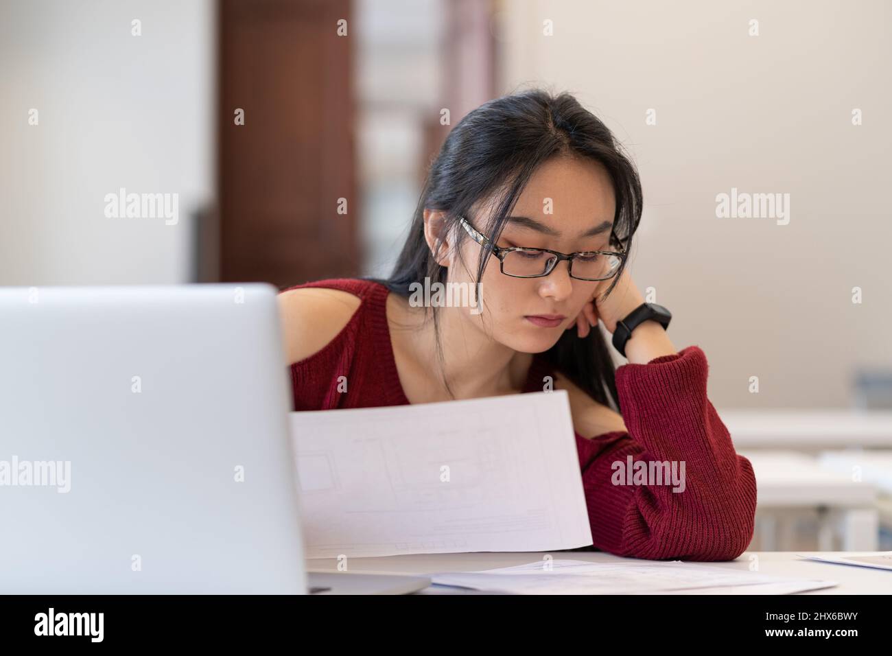 Fleißiges asiatisches Studentenmädchen in Glasen, das während des Studiums in der Bibliothek Architekturzeichnungen liest Stockfoto