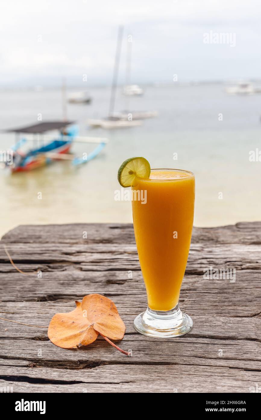 Mango-Saft am Strand mit dem Meer und einem Boot im Hintergrund. Sanur, Bali, Indonesien. Vertikales Bild. Stockfoto
