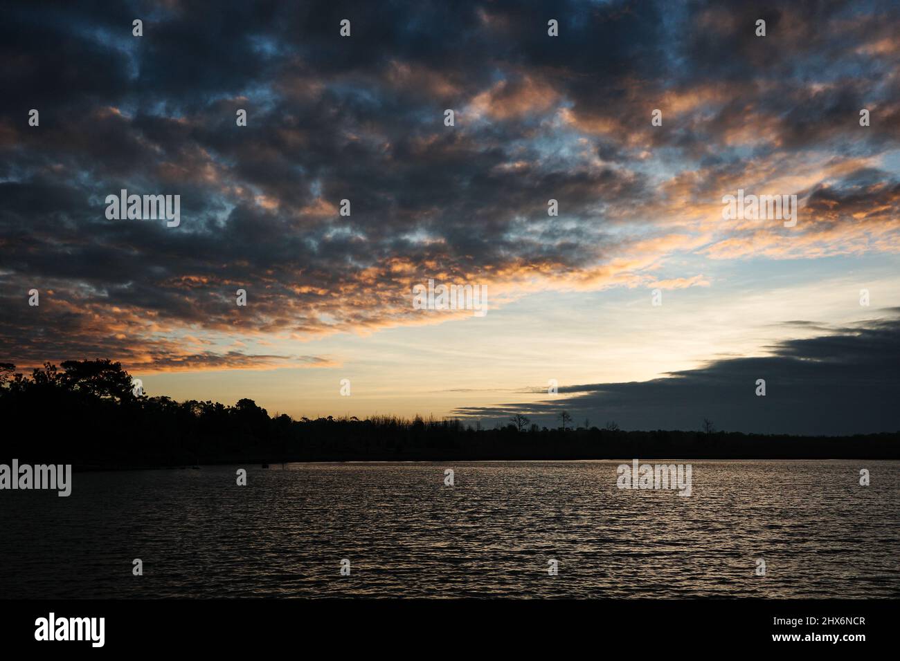 Ruhige Seenlandschaft im dunklen Waldgebiet, der Himmel bedeckt von Wolken, die das goldene Sonnenlicht berühren. Stockfoto