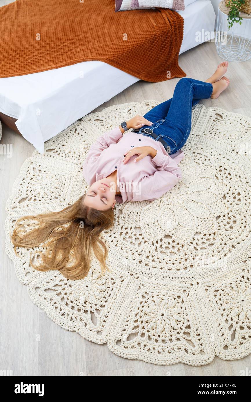 Eine Frau liegt auf einem Teppich in der Nähe des Bettes, ein  selbstgemachter Strickteppich aus dicken Fäden, von Hand gefertigt: Ein  handgefertigter Teppich aus beigen Fäden liegt auf dem Teppich  Stockfotografie -