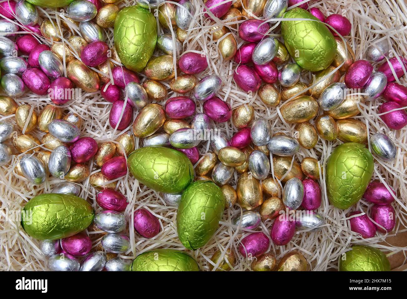 Stapel von bunten Pastellfolie umwickelten Schokoladen-ostereiern in Grün, Rosa, Gelb, Silber und Gold, auf blass gefürchtetem Rahmennest. Stockfoto