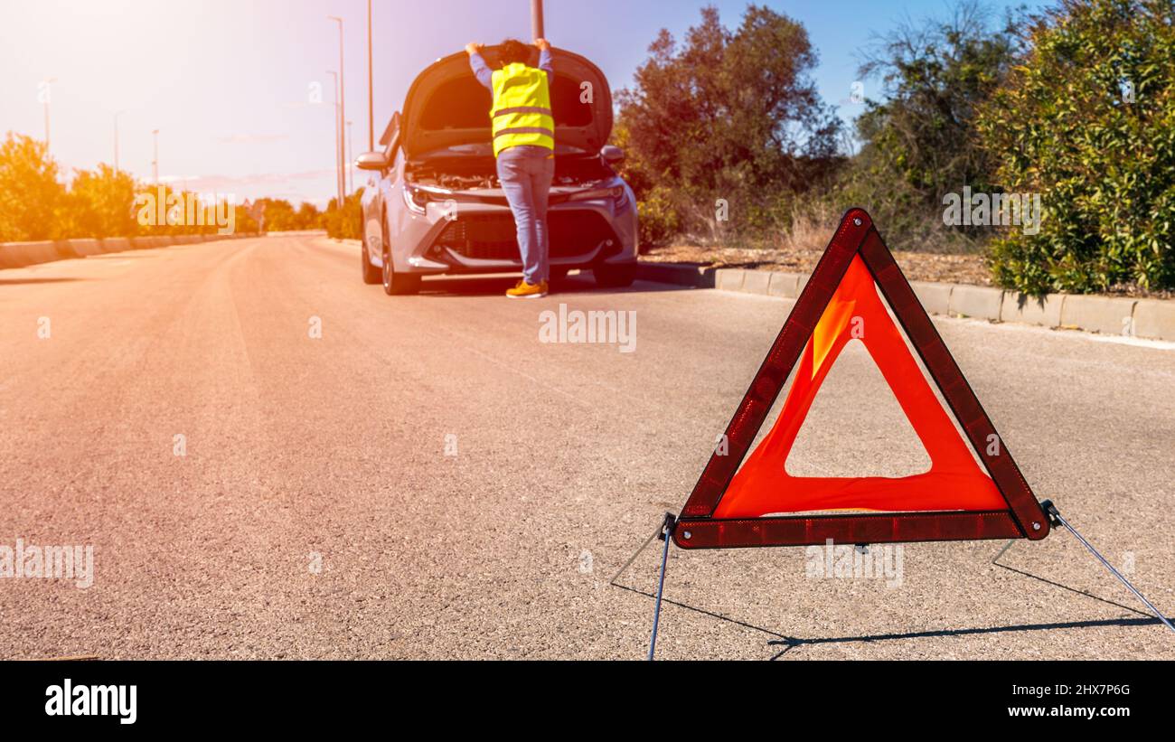 Auto mit Problemen und einem roten Dreieck, um andere Verkehrsteilnehmer zu warnen. Männlicher Fahrer, der in der Nähe eines kaputten Autos mit offener Motorhaube steht. Stockfoto
