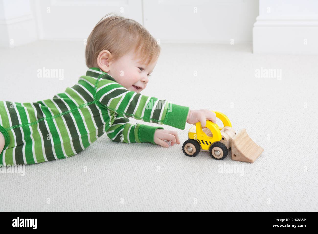 Kleiner Junge, der vorne auf dem Teppichboden liegt und mit einem gelben LKW-Spielzeug spielt Stockfoto