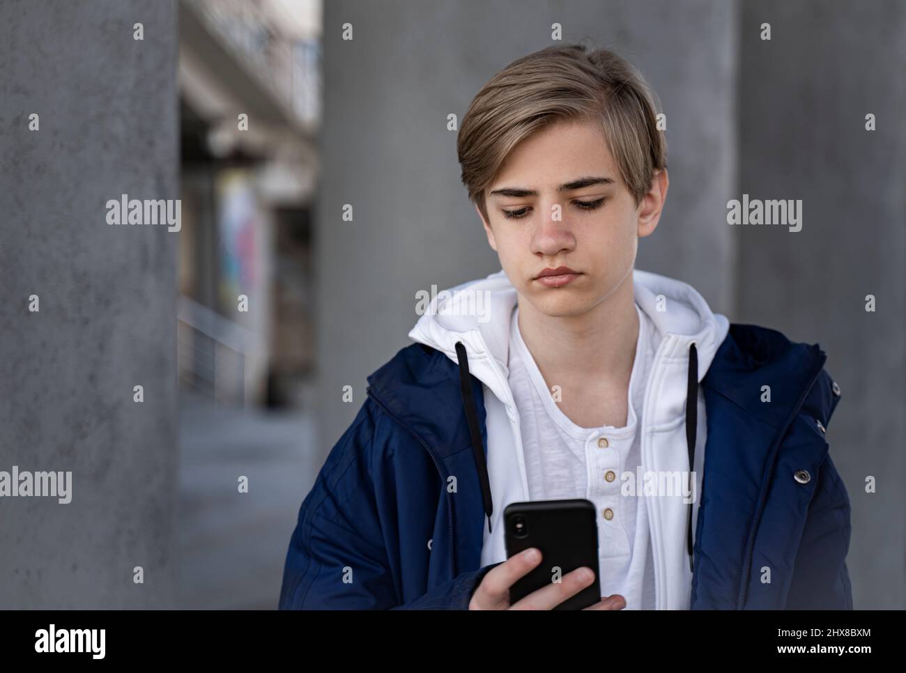 Teenager Junge in einer blauen Jacke und einem weißen T-Shirt hält ein Handy in den Händen, schaut auf die Kamera und lächelt. Stockfoto
