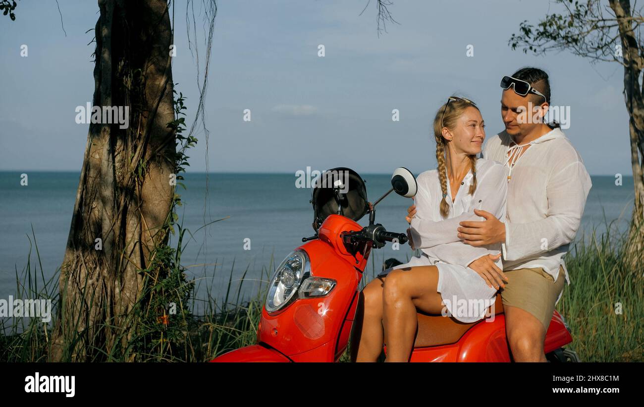 Motorroller Road Trip. Liebespaar auf rotem Motorrad in weißer Kleidung auf Waldweg. Gerade verheiratete Frau und Mann küssen, umarmen, sitzen auf dem Motorrad. Weddin Stockfoto