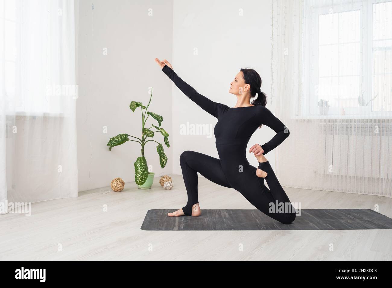 Frau in schwarzer Sportkleidung, die Yoga praktiziert und mit virasana anjaneyasana trainiert, in niedriger Ausfallhaltung, sitzend auf einer Matte in einem Raum am Fenster Stockfoto